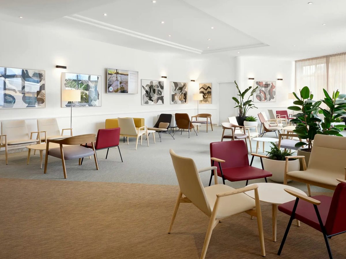 An indoor lounge with colourful armchairs, coffee tables, wall art and potted plants by large windows.