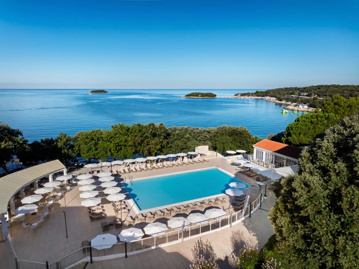 A large rectangular freshwater pool ringed with white parasols and loungers on a terrace above the sea, with wooded islets offshore.