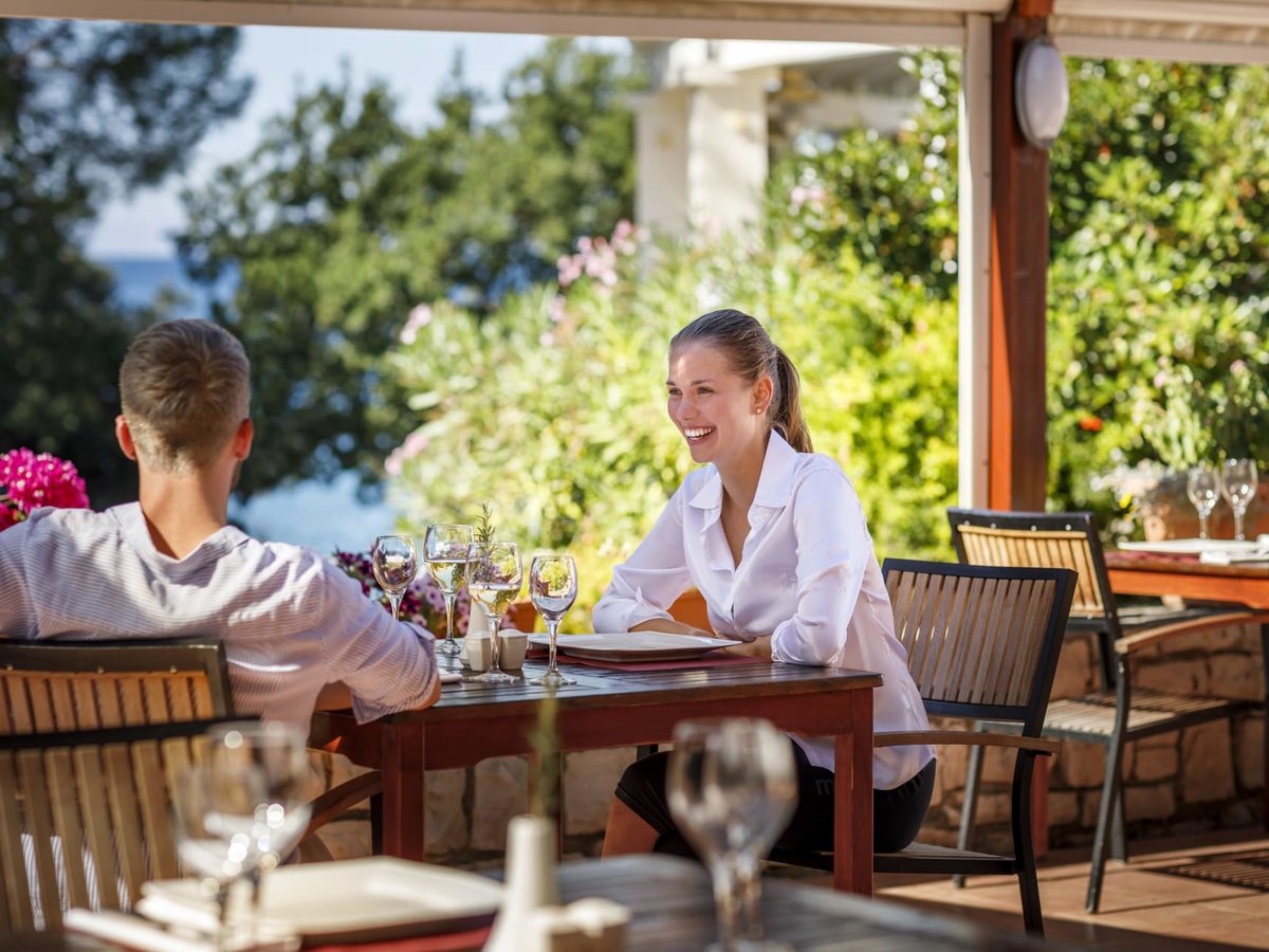 Two diners at a laid table with wine glasses on a covered garden terrace, with flowering shrubs and a glimpse of the sea beyond.