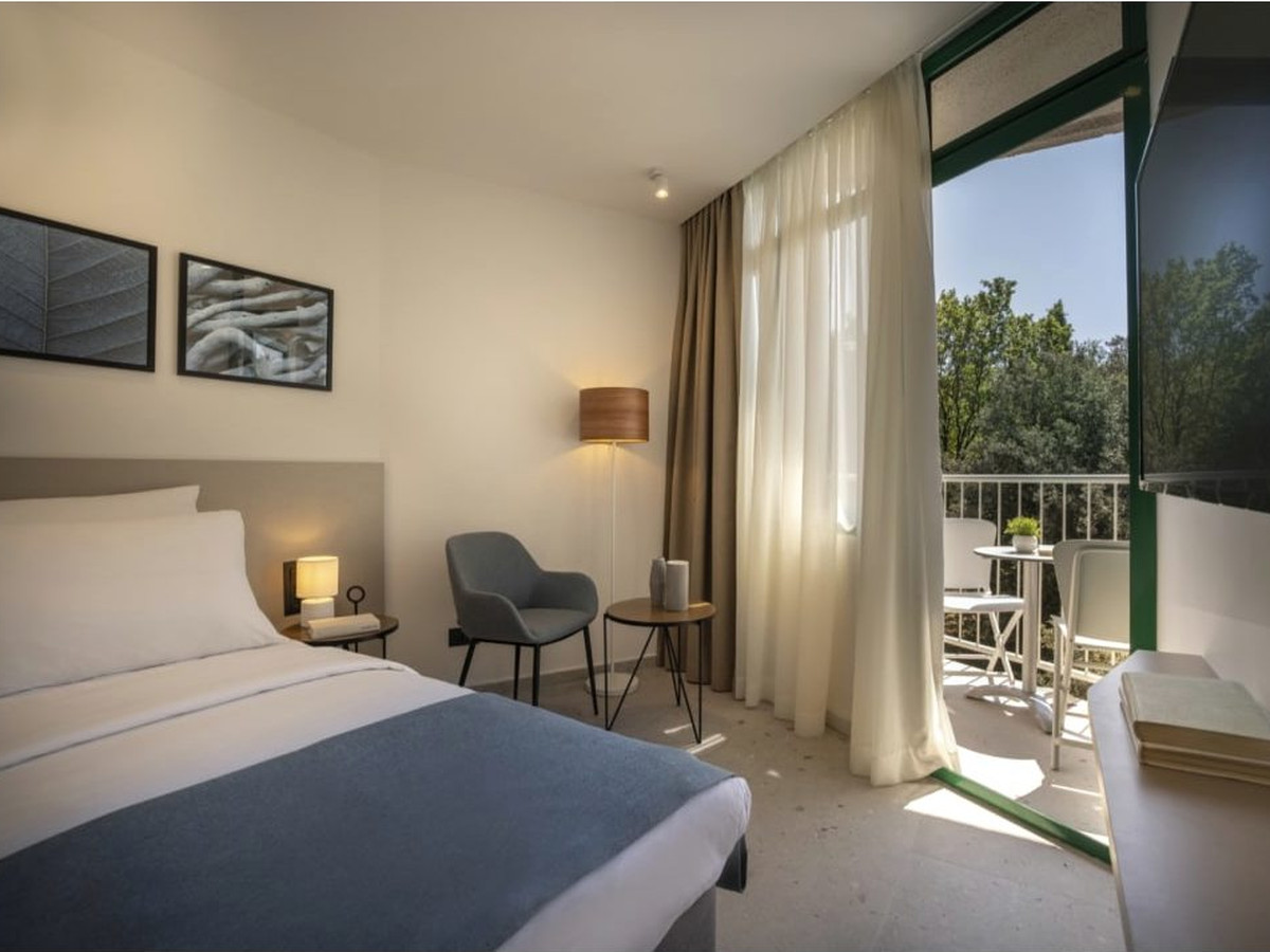 A double room with a double bed, a blue bed runner, a grey armchair and a balcony door onto trees.