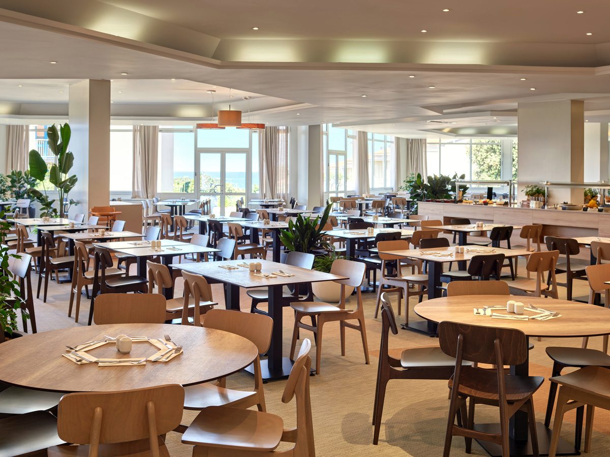An indoor buffet restaurant with round and rectangular wooden tables under a coffered ceiling with recessed lights and large windows onto greenery.