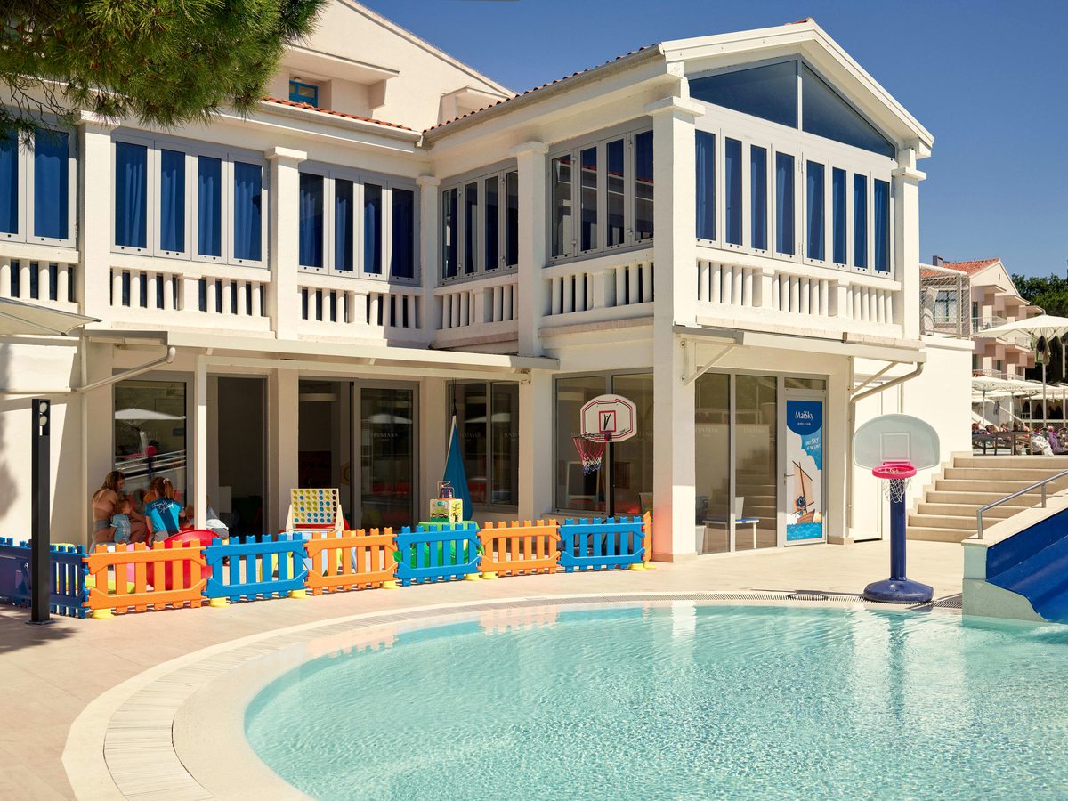 A shallow curved children's pool edged with low colourful fencing in front of the white Mini Club building.