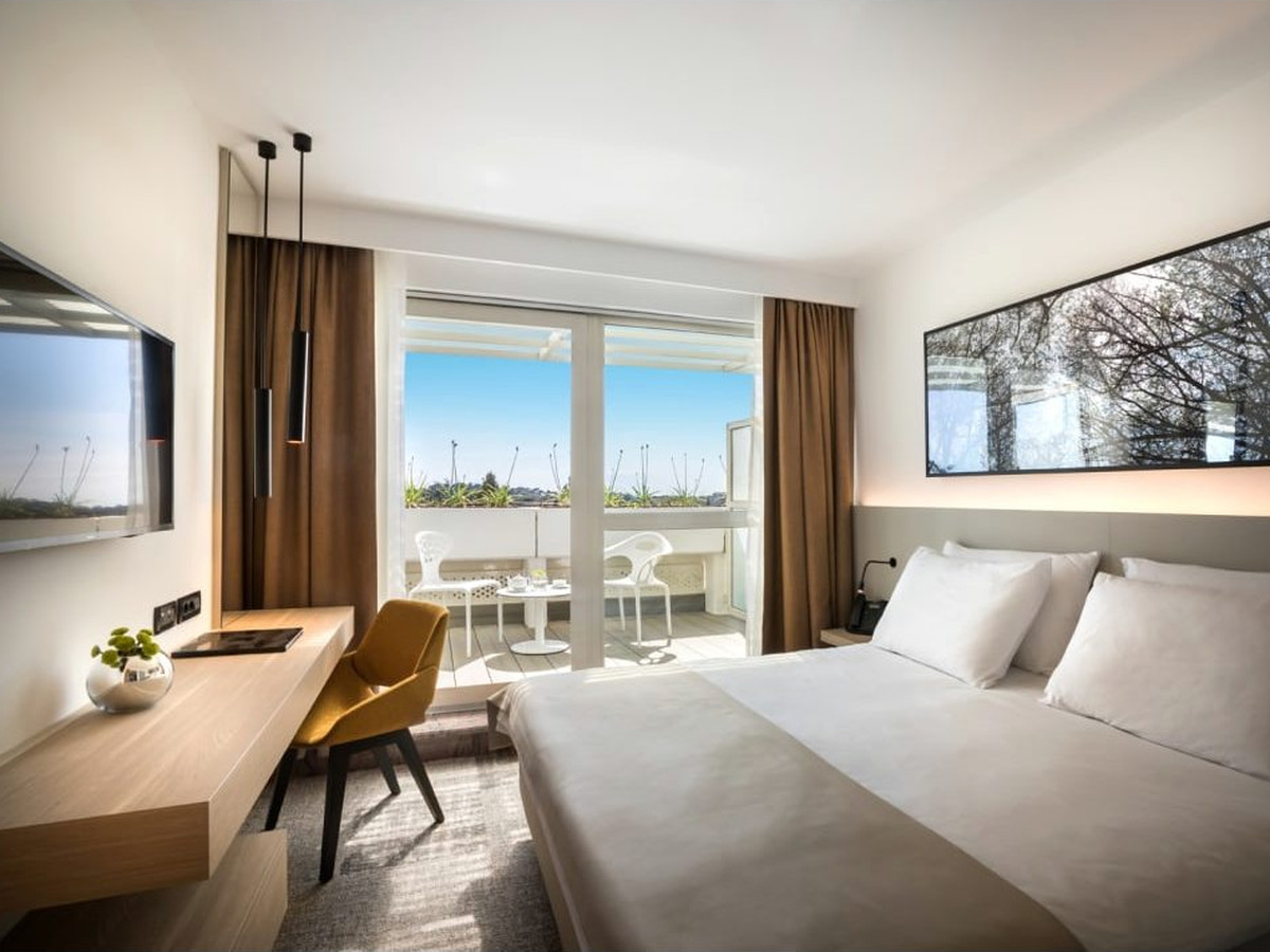A double room with a writing desk, an armchair and a balcony with a table and chairs looking onto trees.