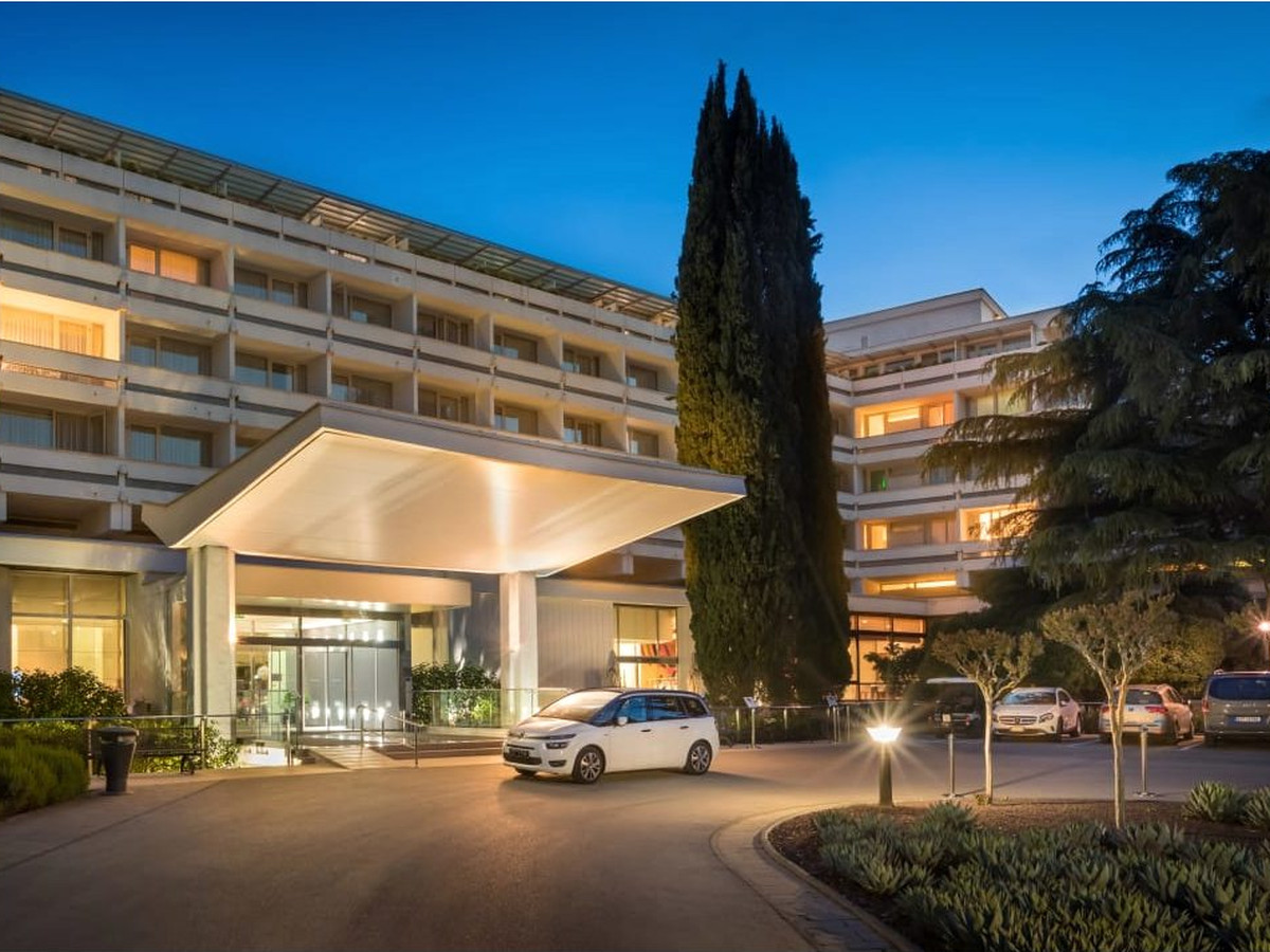 The resort's main entrance building, with a canopy over the drop-off, a car parked beneath it and cypress and pine around it.