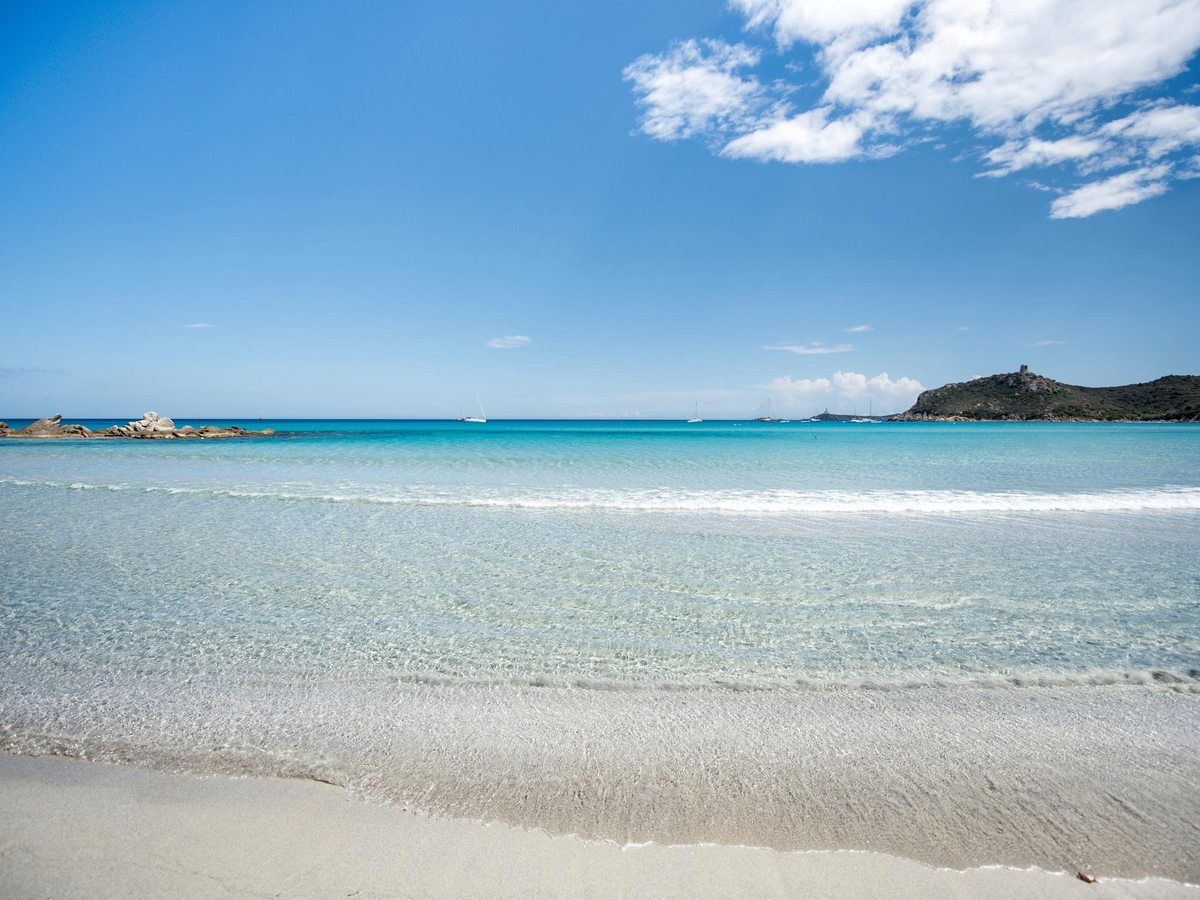 A wide white-sand beach meeting clear shallow turquoise water, with a rocky headland at one end under a blue sky.