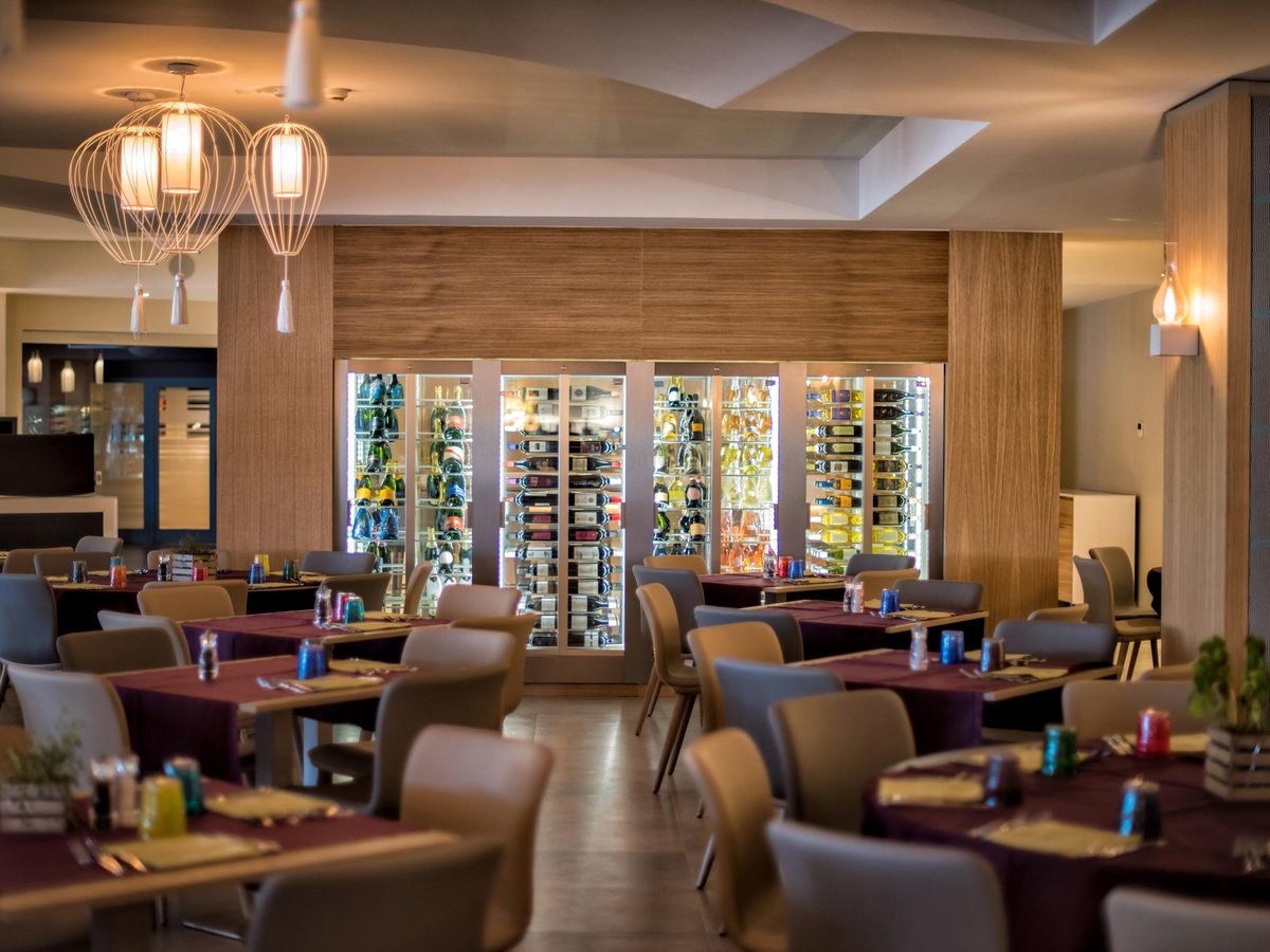 Set dining tables with upholstered chairs in a warm-lit indoor restaurant, a wall of glass-fronted wine fridges behind.