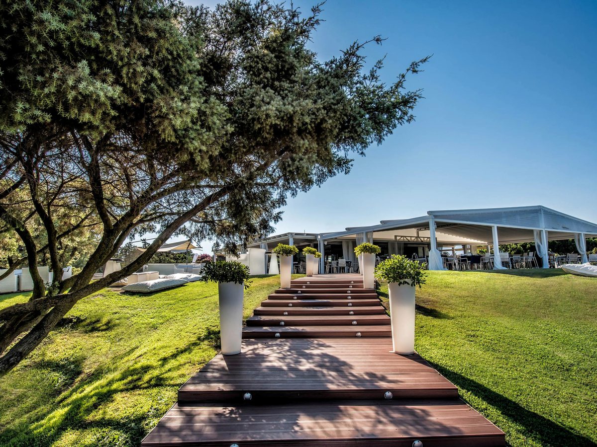 A wooden boardwalk path climbing across landscaped lawns to an open white pavilion, with a large juniper tree overhanging and hills behind.