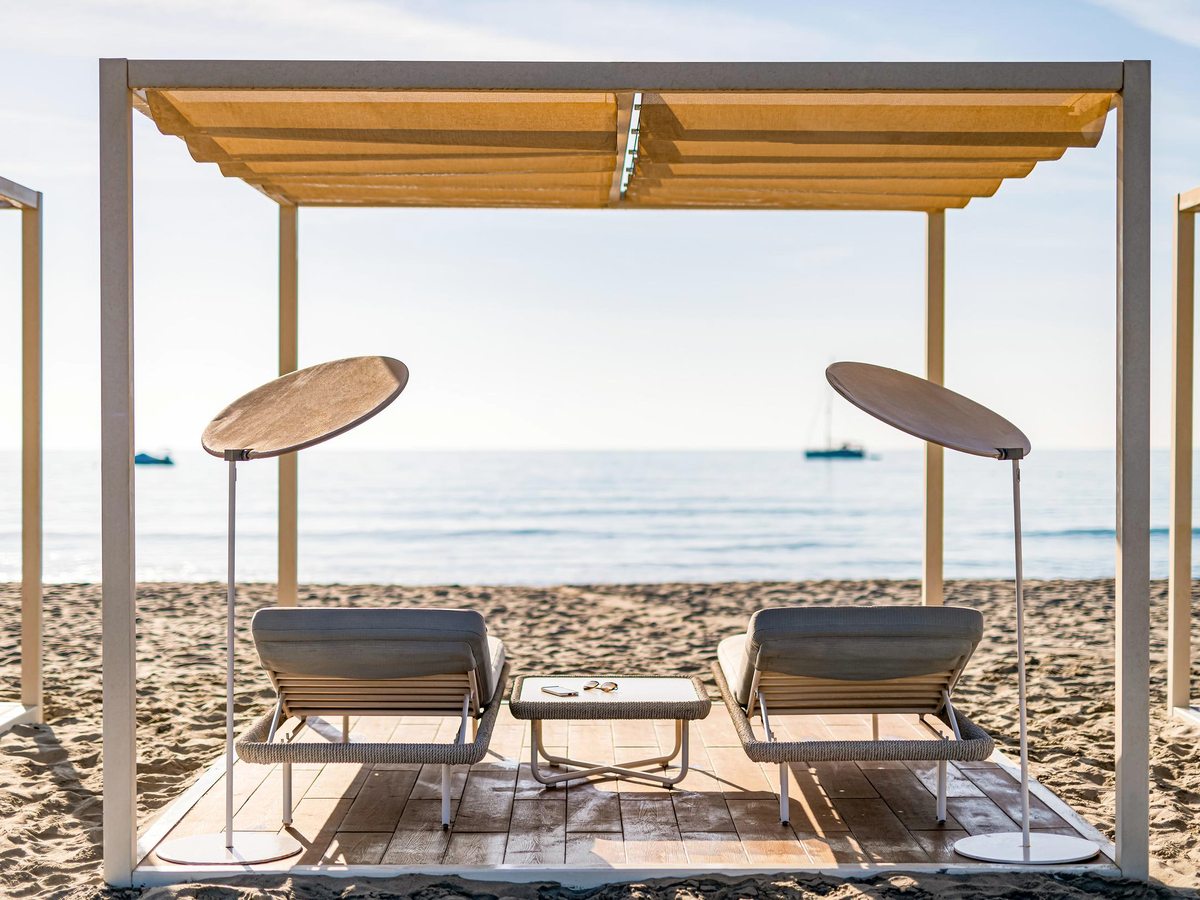 A wooden pergola shading two sun loungers and a small table on the sand, facing the calm sea with boats offshore.