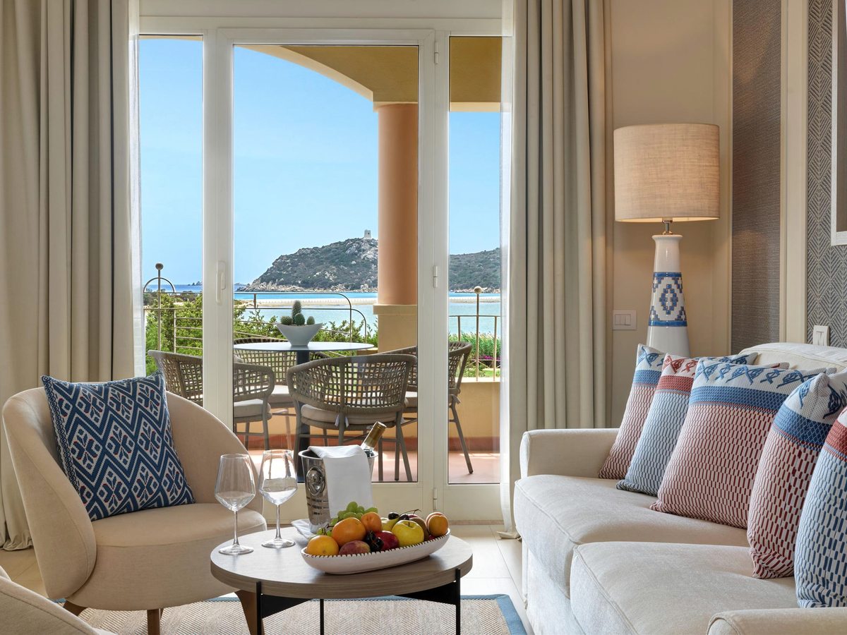 A suite lounge with a white sofa, armchairs and a coffee table, floor-to-ceiling doors opening to a terrace above a turquoise bay and headland.