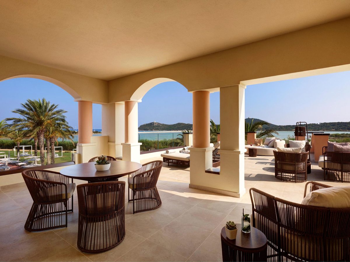 A shaded suite terrace with an outdoor dining table, wicker chairs and lounge seating framed by arches, with palms and the sea beyond.