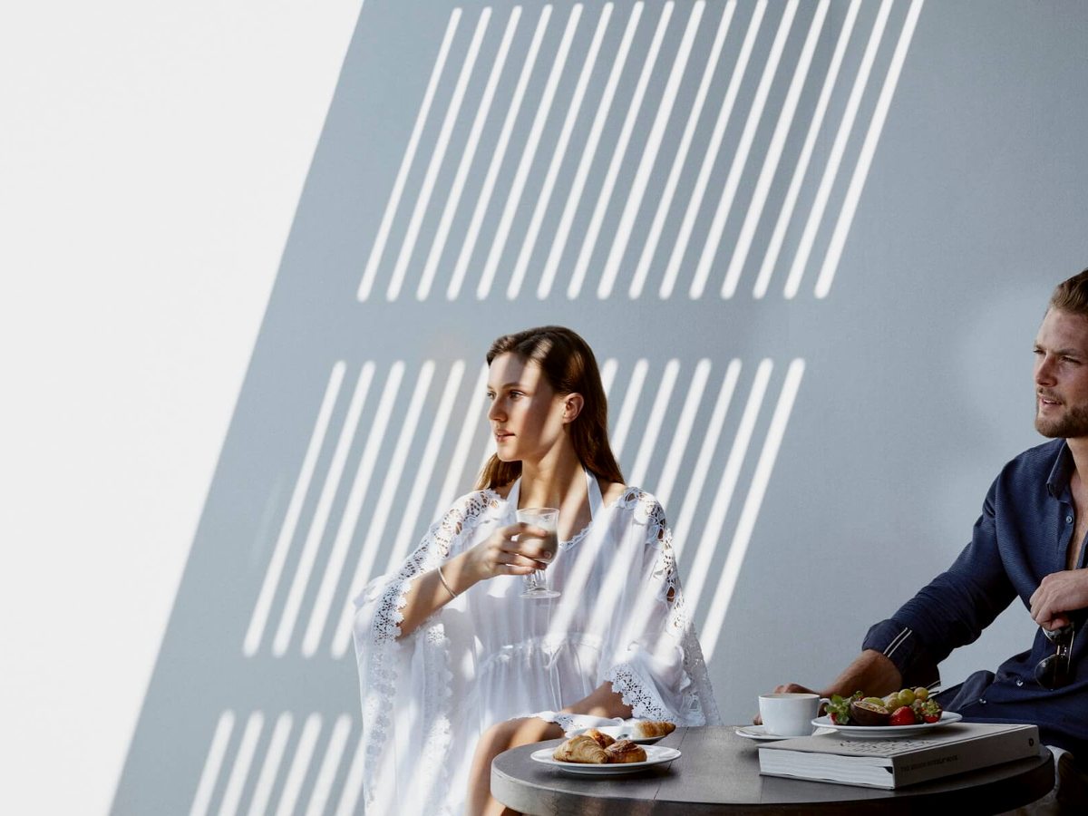 A couple at a small round table in the sun with a plate of croissants, fresh fruit and coffee, striped light falling on the wall behind.