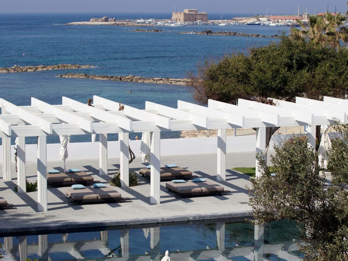 White pergola-shaded daybeds on a stone terrace beside a pool, with Paphos Harbour, the marina and the medieval castle across the water.