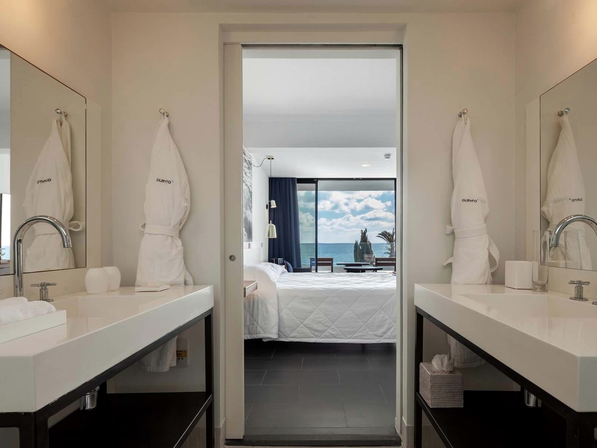 A bathroom with two white basins on black stands and Almyra robes, a sliding door open to a sea-view bedroom beyond.