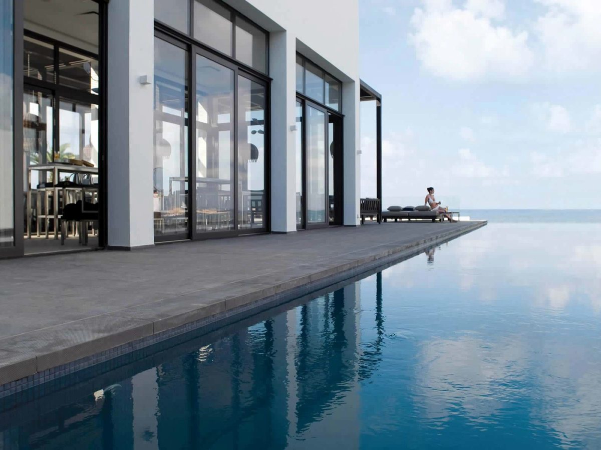 A long infinity pool running beside a glass-fronted terrace to a seamless edge over the sea, a single guest seated at the far end.