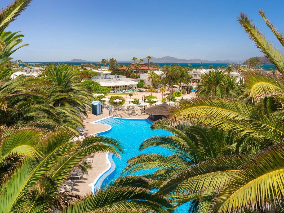 An elevated view over the resort's low white buildings, palm gardens and main pool, with the Atlantic and distant islands on the horizon.