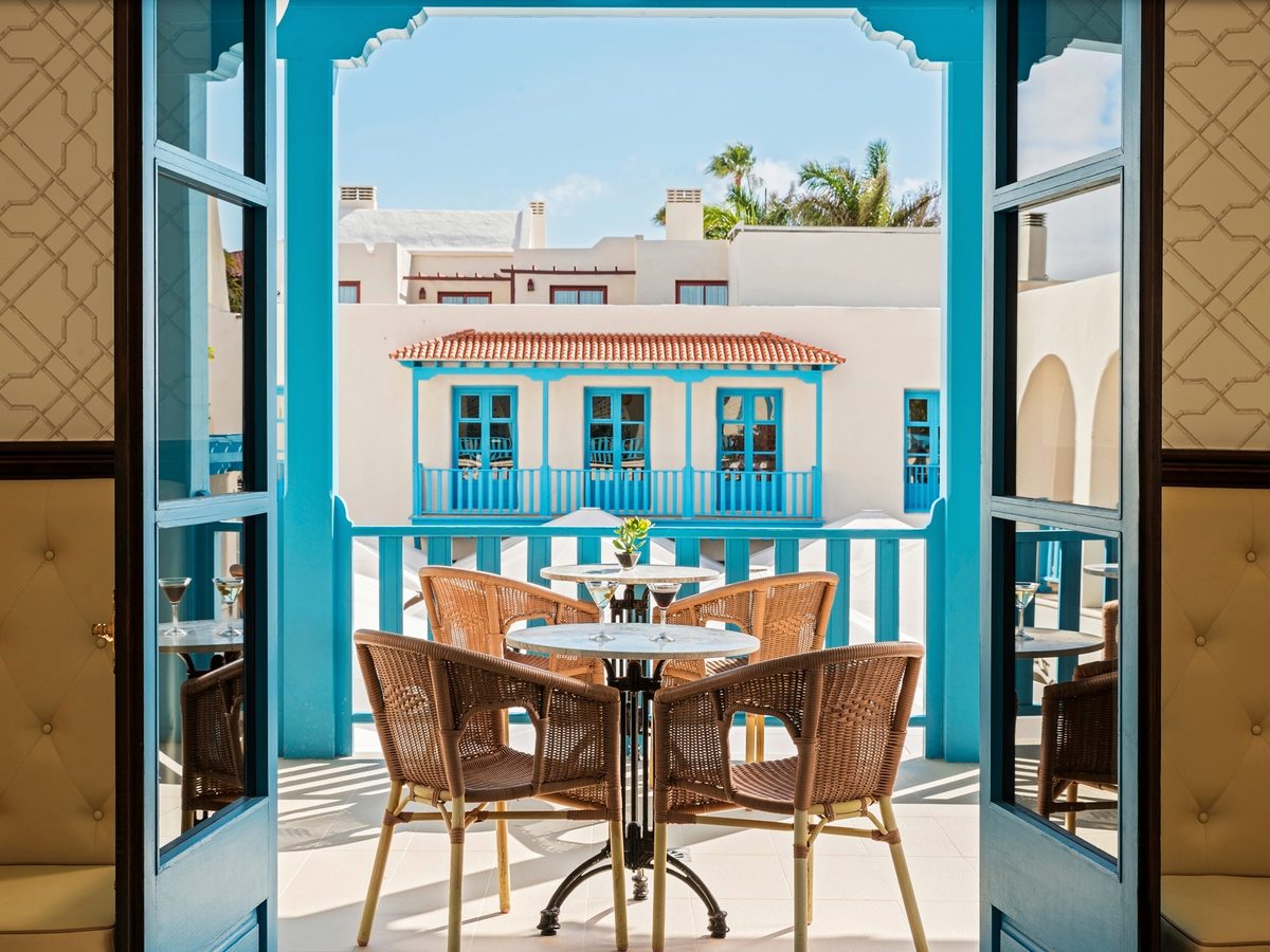 An indoor lounge with cushioned wicker chairs at marble-topped bistro tables and upholstered banquettes, opening through blue louvred doors onto the resort's whitewashed, blue-trimmed buildings.