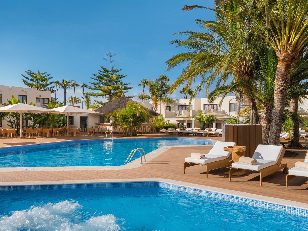 A curved pool beside a thatched-roof poolside bar, with cushioned loungers on the deck, tall palms and a bubbling whirlpool in the foreground.