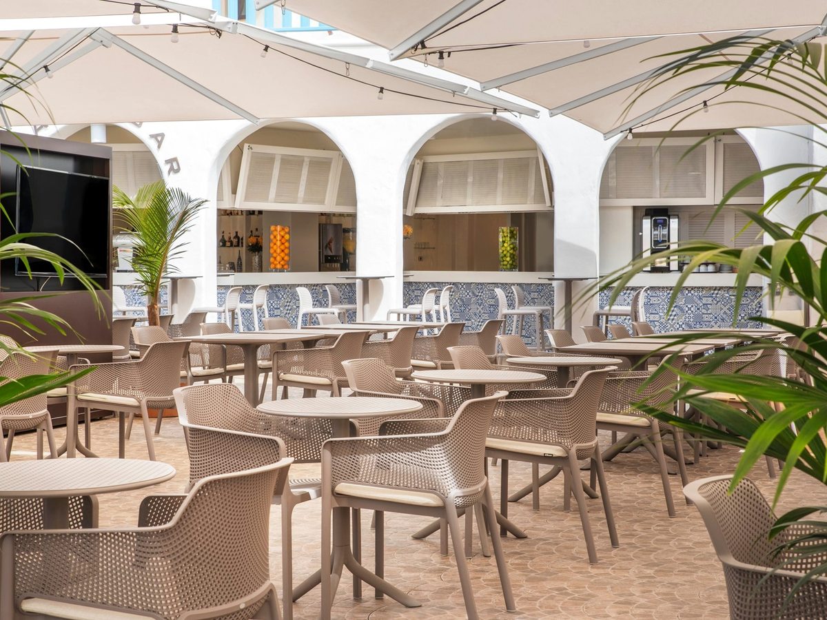An outdoor bar terrace with cafe tables, woven chairs and large parasols under a white arched facade.
