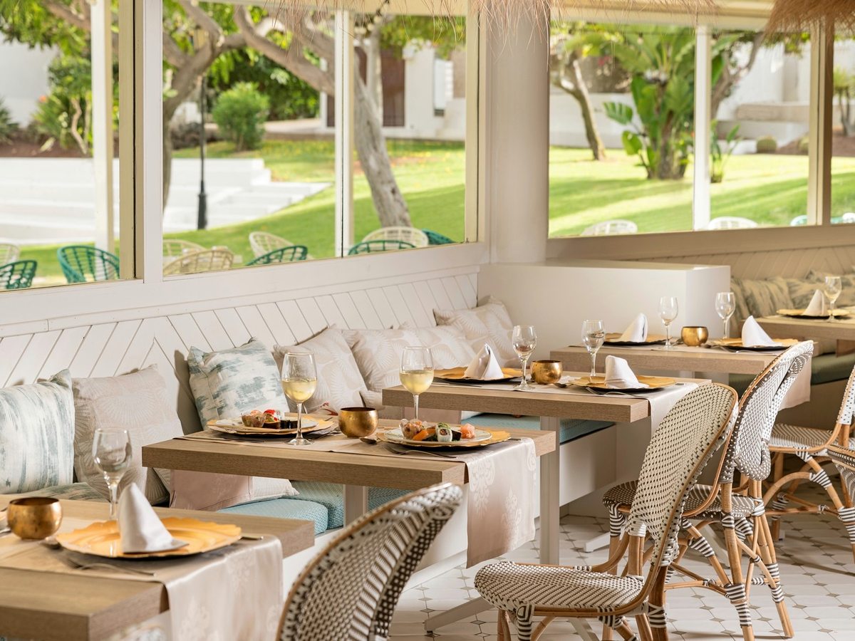 A bright dining room with laid tables, bench seating and cane chairs beside full-height windows onto the lawns.