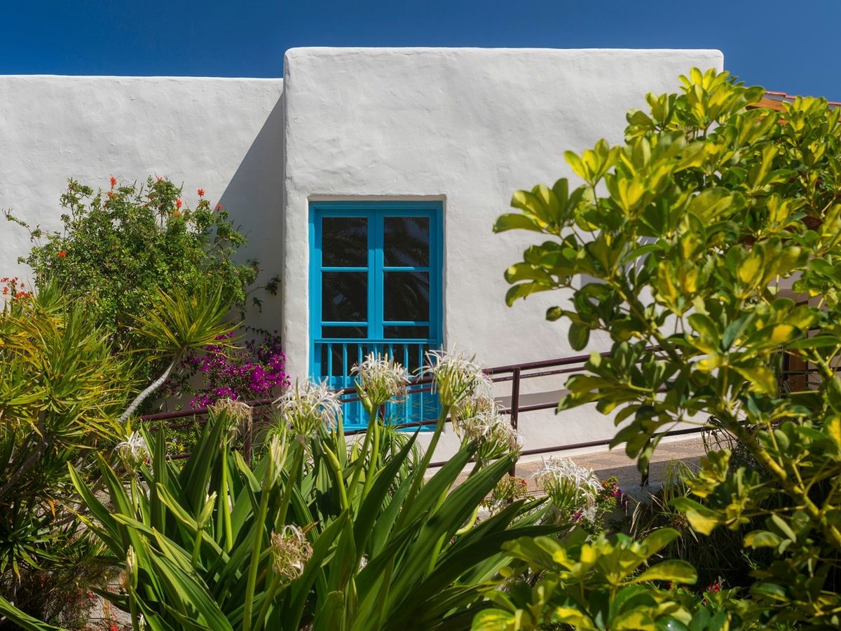 A whitewashed suite building with a bright blue door and window, backed by flowering shrubs and pink bougainvillea along a garden path.