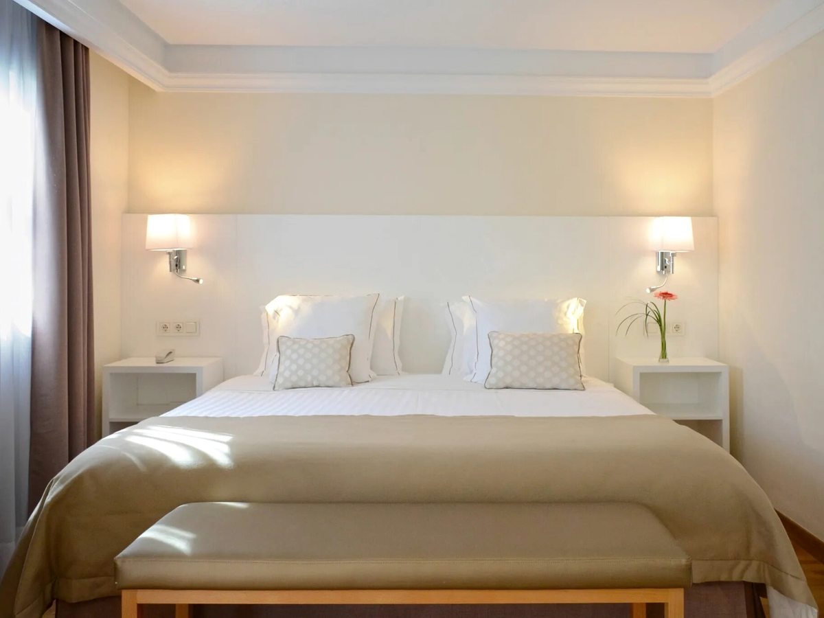 A suite bedroom with a large double bed, a padded headboard, matching bedside tables and wall lamps in neutral tones.