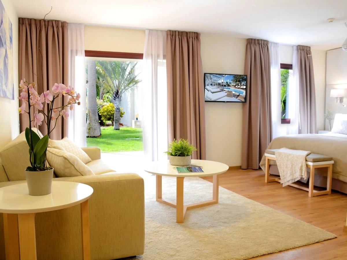 A suite living area with a sofa and coffee table and sliding glass doors opening onto a garden lawn with palms, a bed to one side.