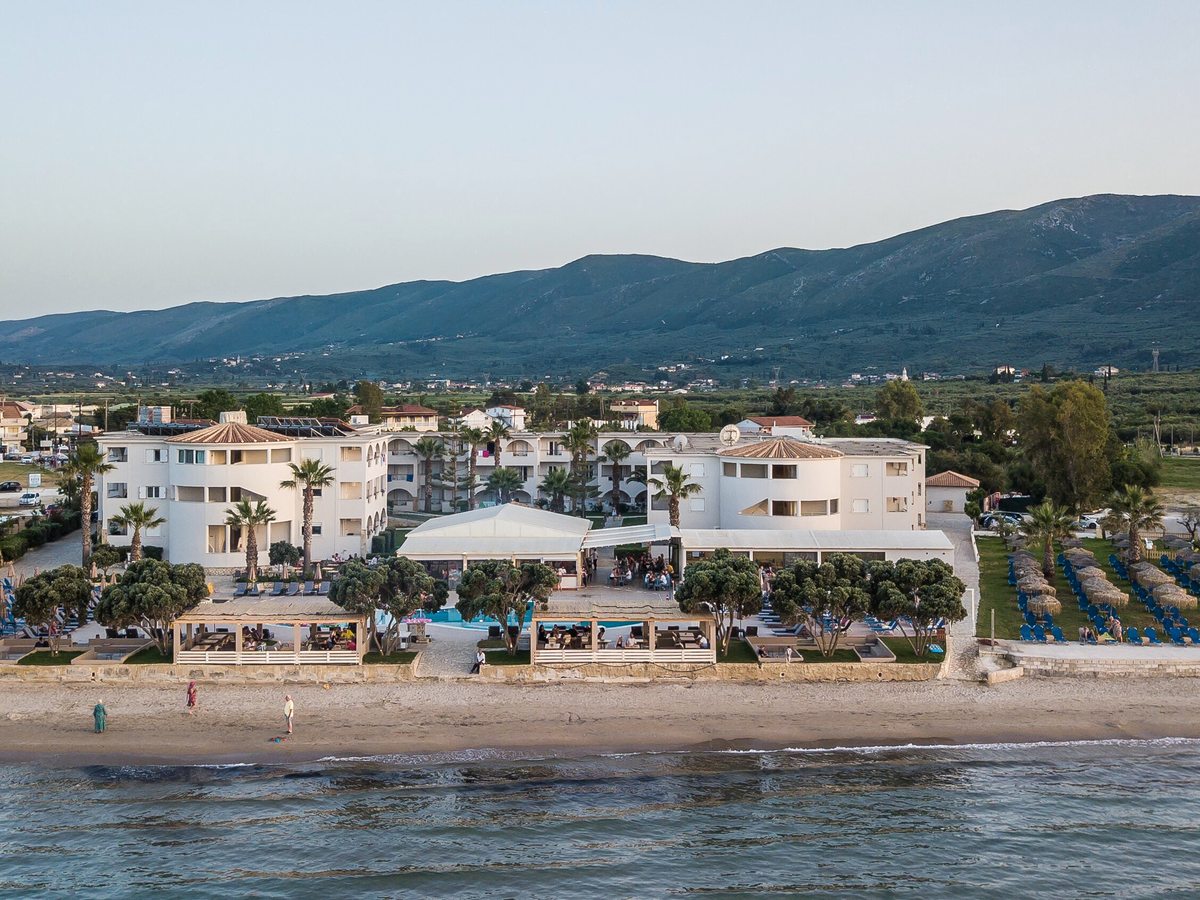 Aerial view of the white low-rise resort buildings stretched along the sandy Alykanas beachfront, with green hills rising behind under a clear sky.