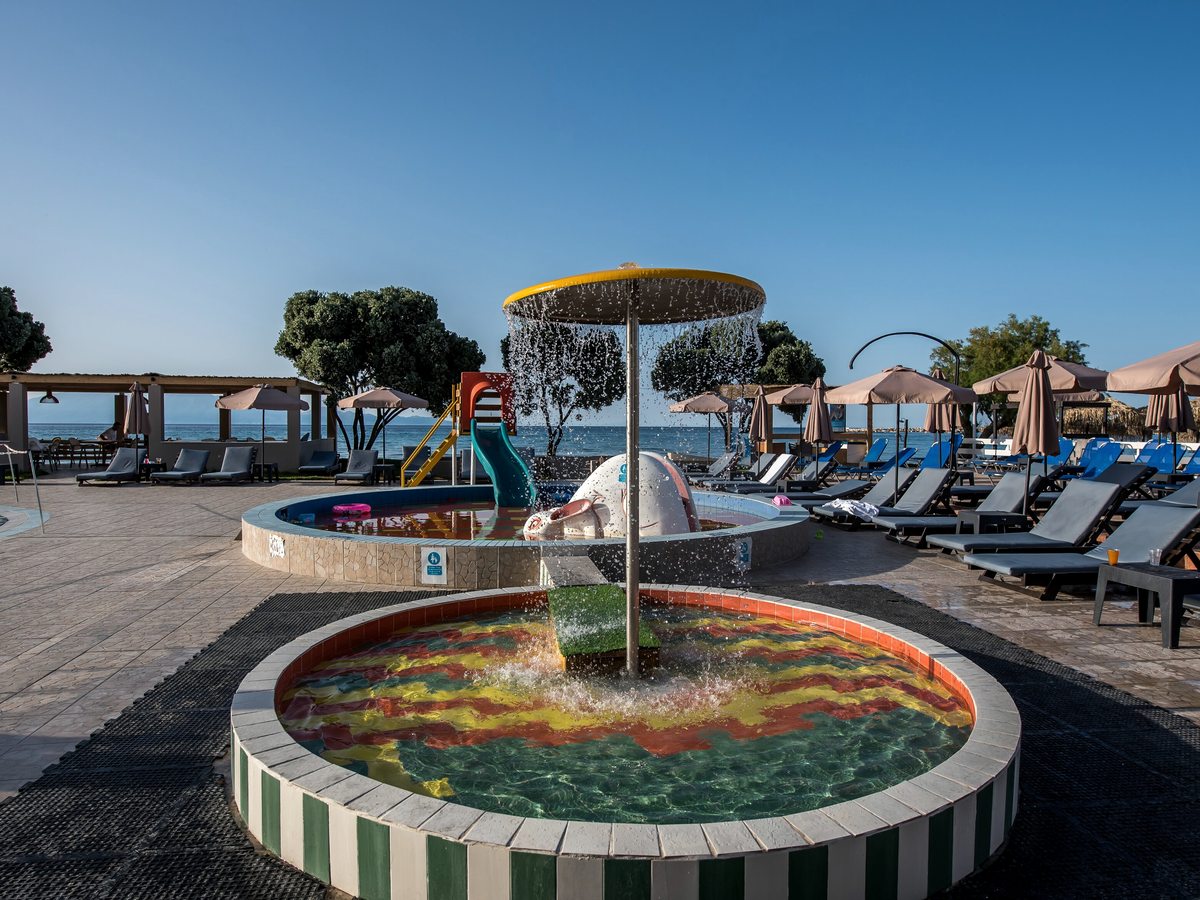 Circular shallow children's pool with a mushroom fountain and a small slide, sunbeds and the sea behind.