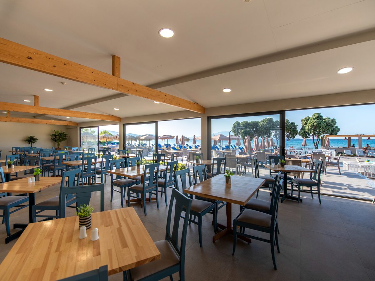 Restaurant dining room with wooden tables and blue chairs beside large windows and sliding doors opening to a terrace and the sea.