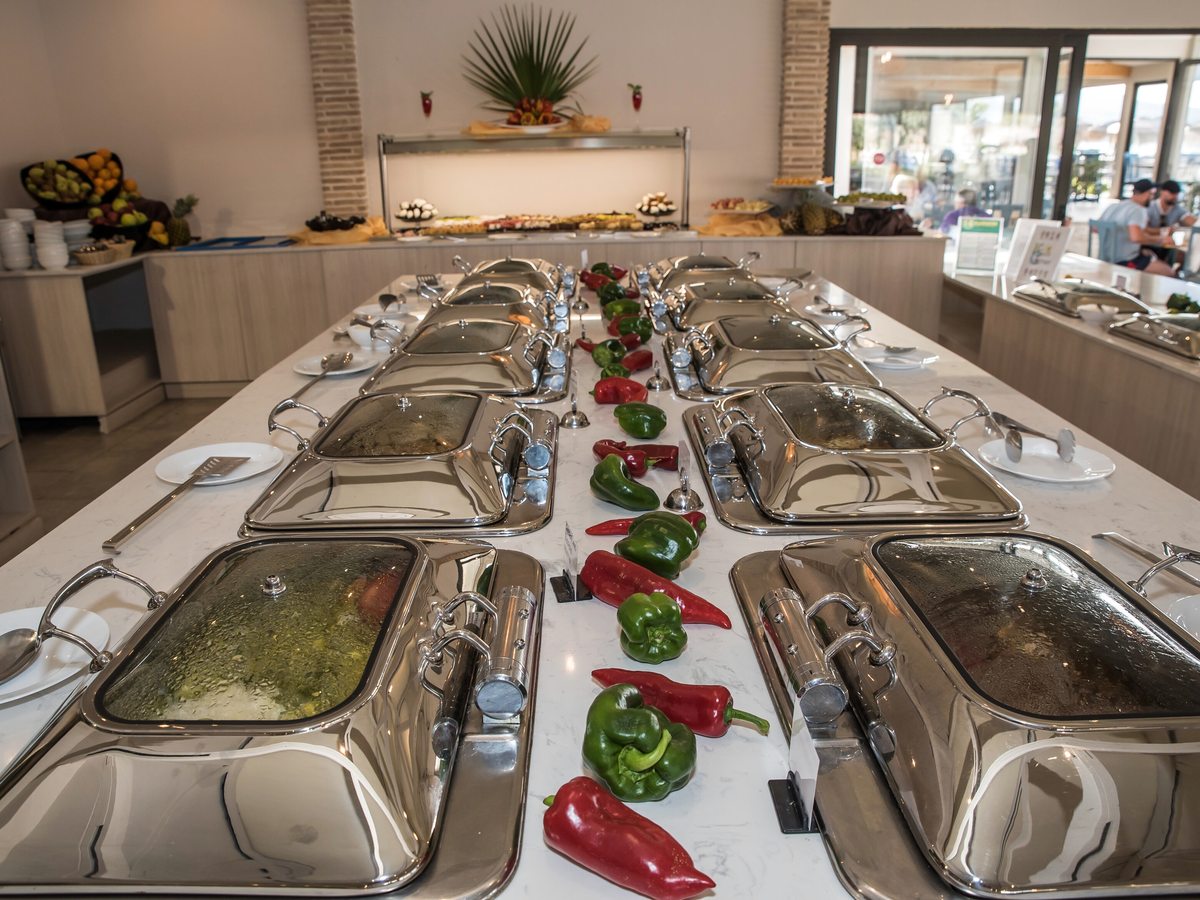 Long buffet island of stainless-steel chafing dishes with hot food, garnished with red and green peppers.