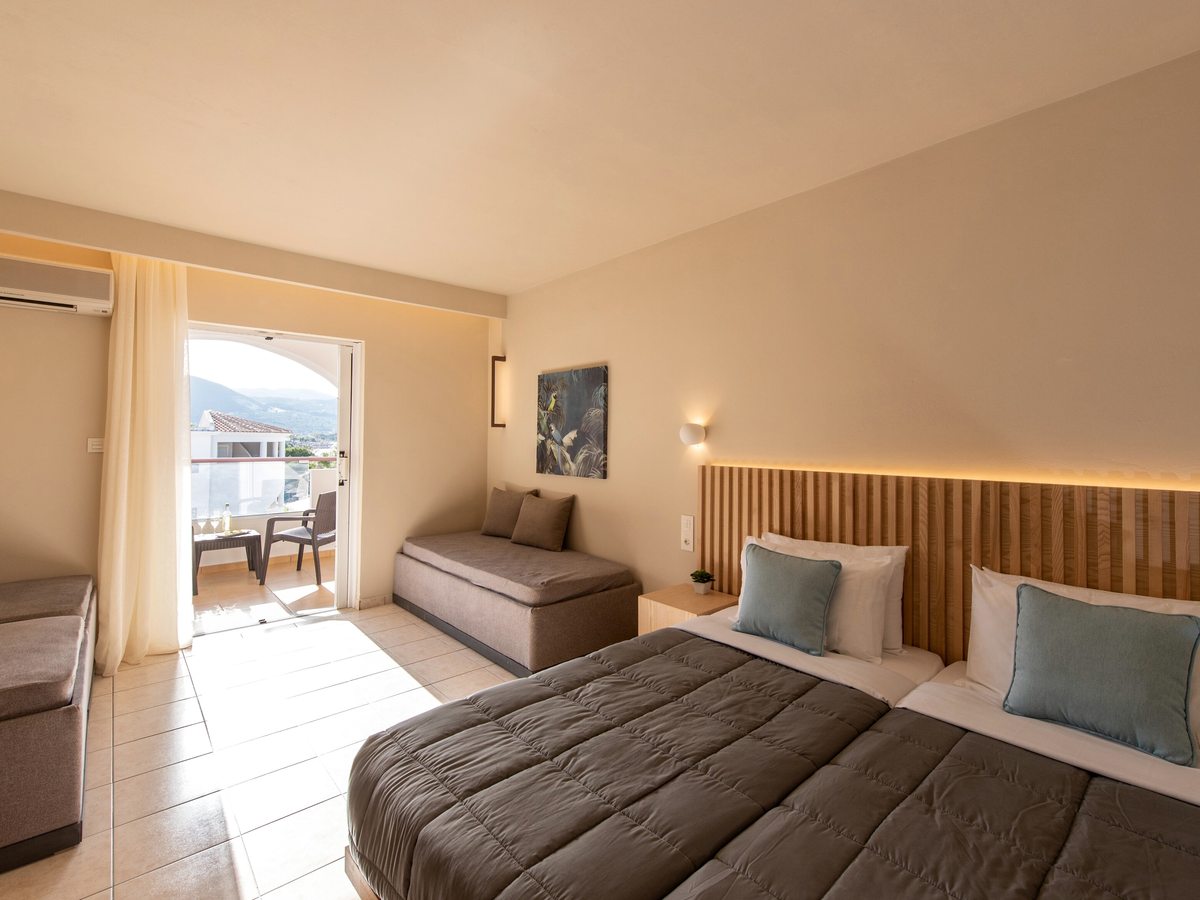 Double room with a grey-quilted bed under a backlit slatted-wood headboard and blue cushions, a daybed to one side and a balcony with a table and chairs looking out over rooftops to green hills.