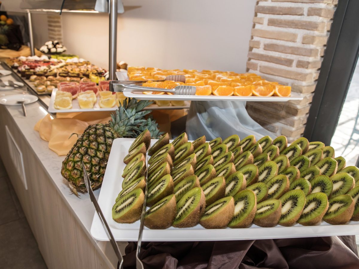 Buffet counter with sliced kiwi, oranges and pineapple, pastries and cold meats on a white service island.