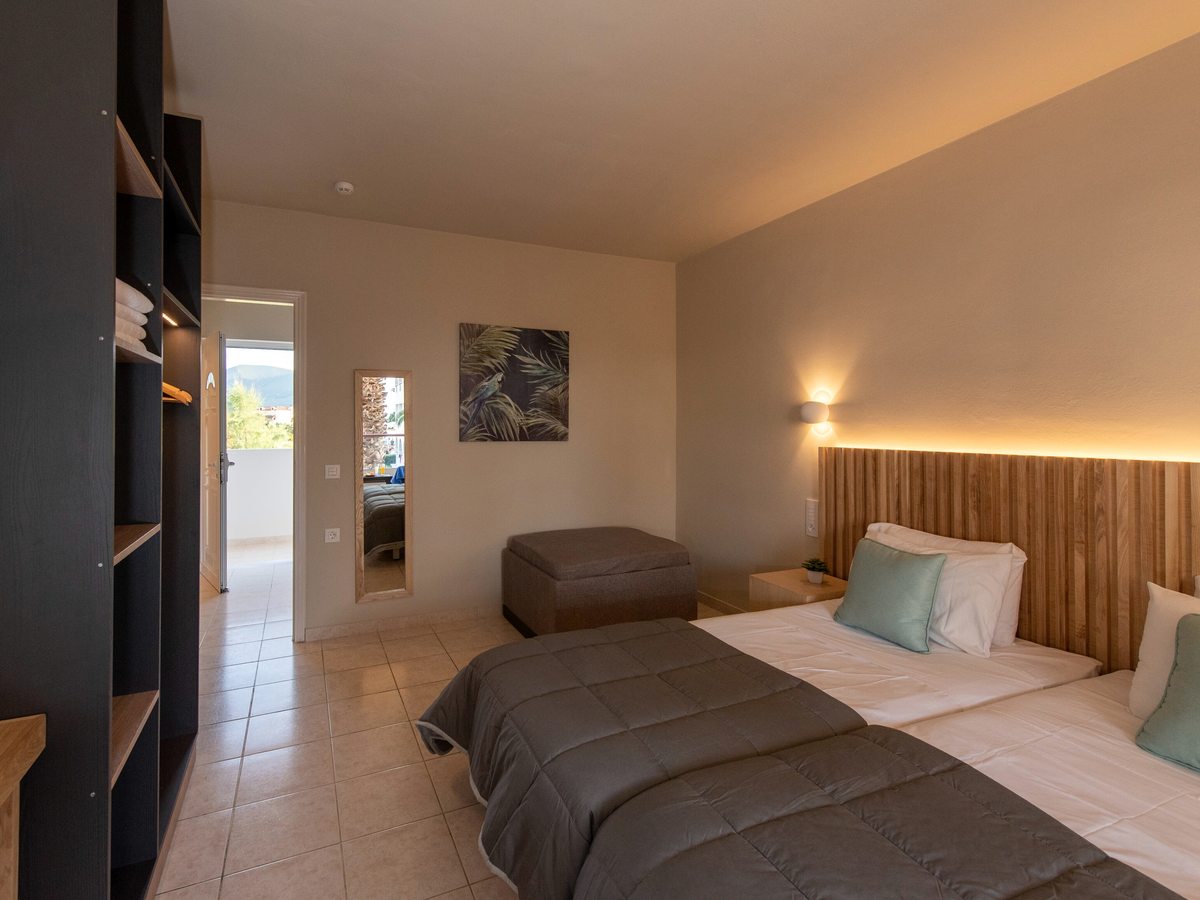 Room with two single beds under illuminated slatted headboards, an ottoman bench at the foot and an open wardrobe to one side.
