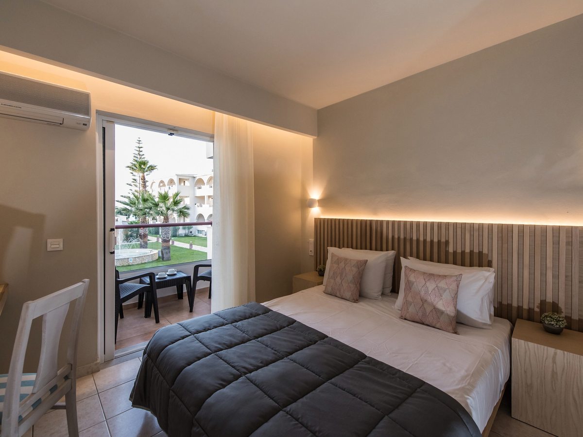 Double room with a slatted headboard and sliding doors opening onto a furnished balcony with a lawn, palm trees and resort buildings beyond.