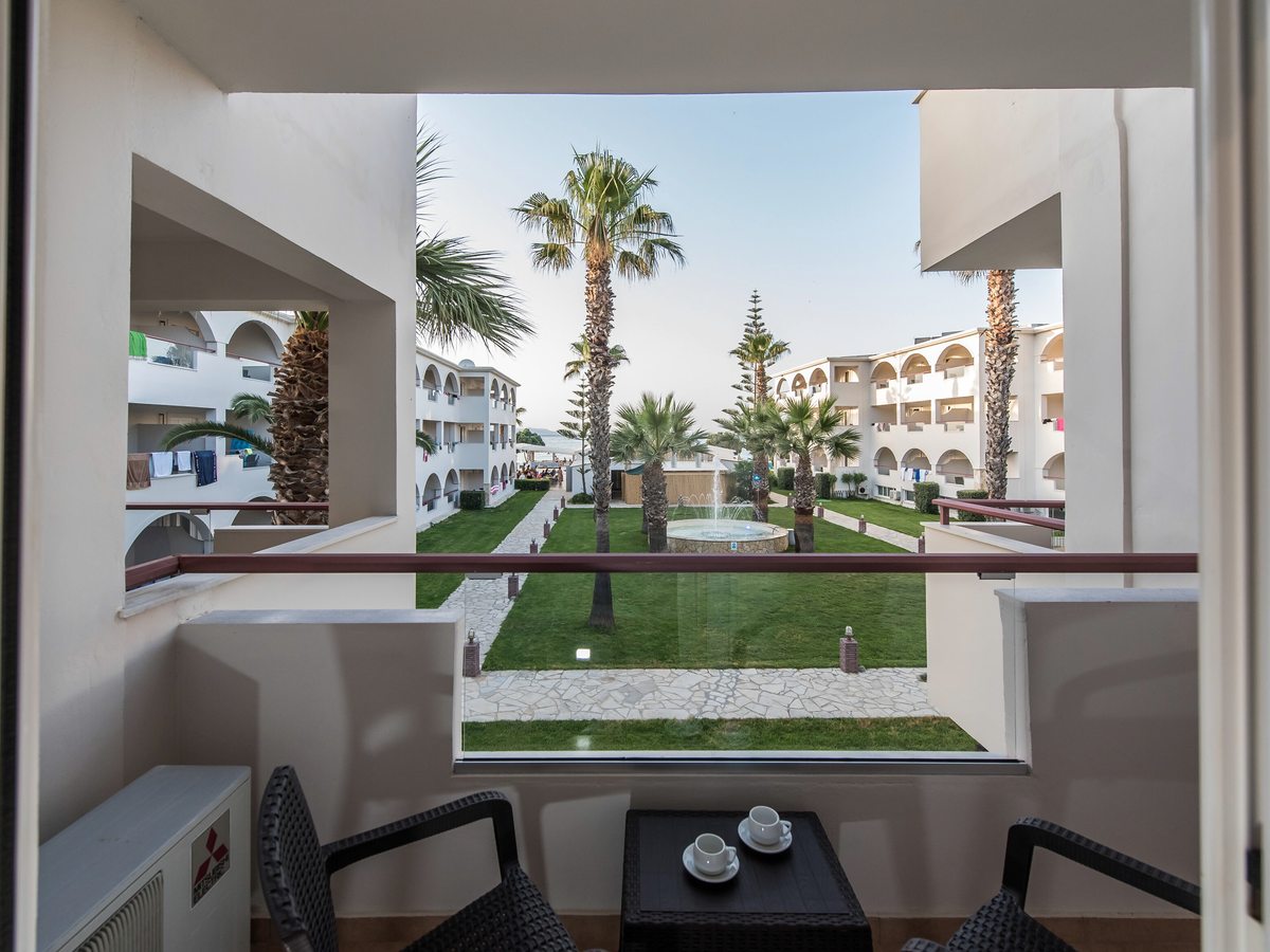 View over a landscaped garden courtyard with palm trees and lawns, framed by white hotel blocks with arched balconies.