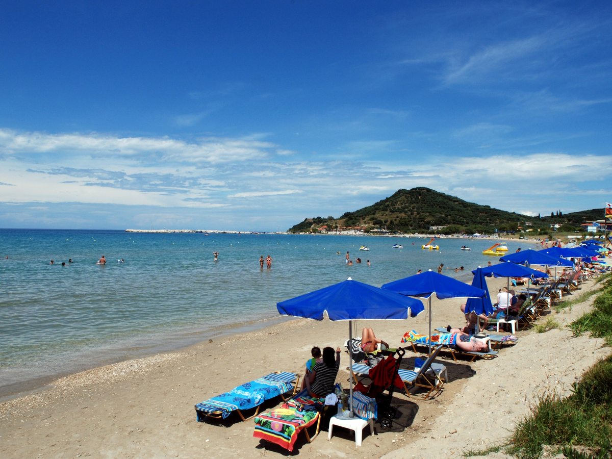 Sandy beach lined with blue parasols and sunbeds beside calm shallow sea, with a green headland in the distance.