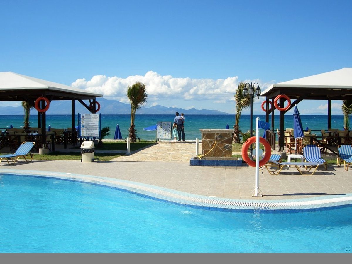 Curved outdoor pool with blue sunloungers and a shaded pavilion beside it, the sea and a headland behind.