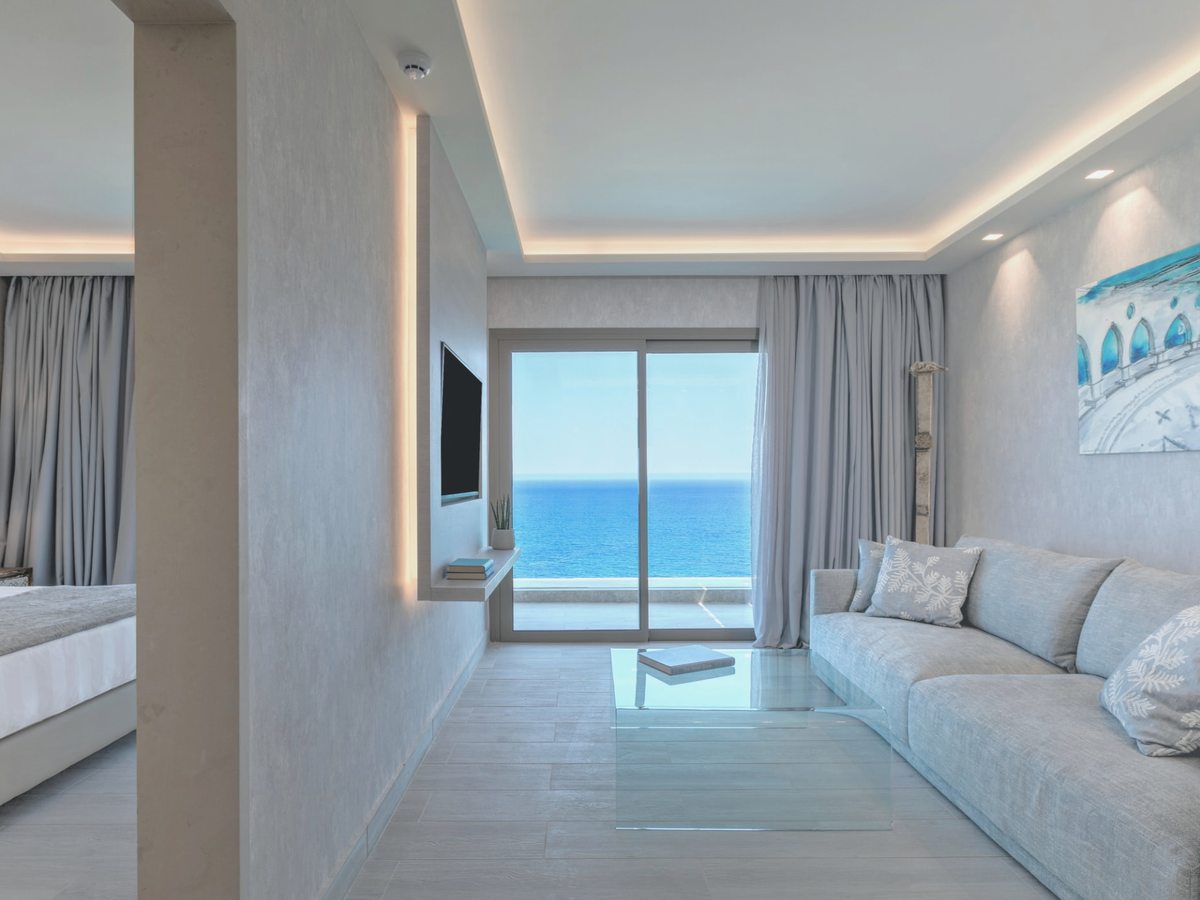 A bright suite with a grey sofa and glass coffee table, a wall-mounted TV and a partition, the bedroom to the left and a sea view through sliding doors.