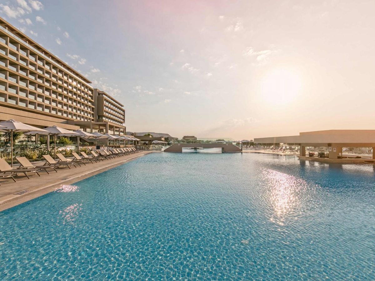 A long turquoise pool running alongside the resort building, lined with sunloungers and parasols under a bright hazy sky.