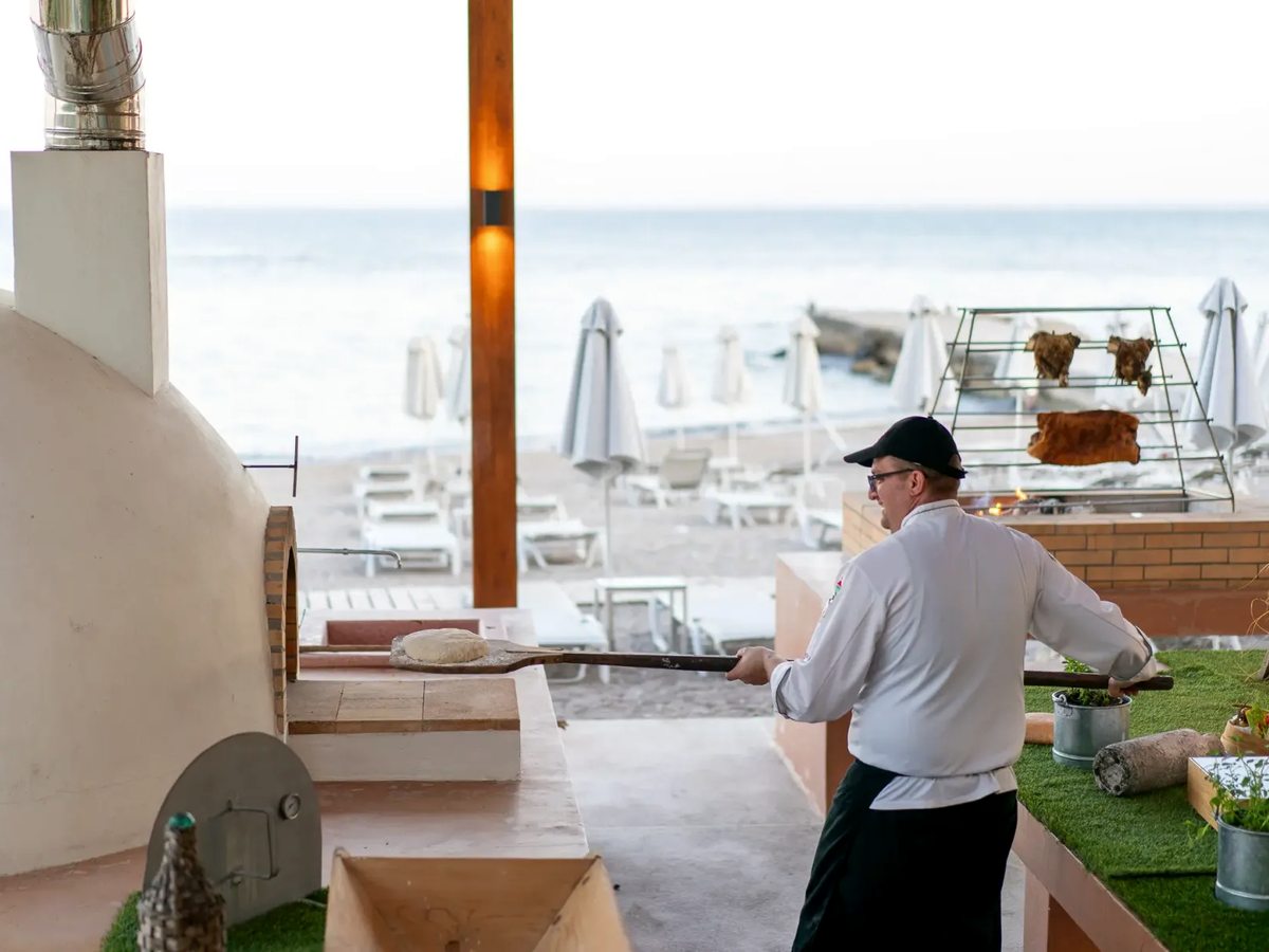 A chef slides dough into a beachfront oven while cuts of meat roast on a rack over an open fire, with sunloungers and the sea beyond.