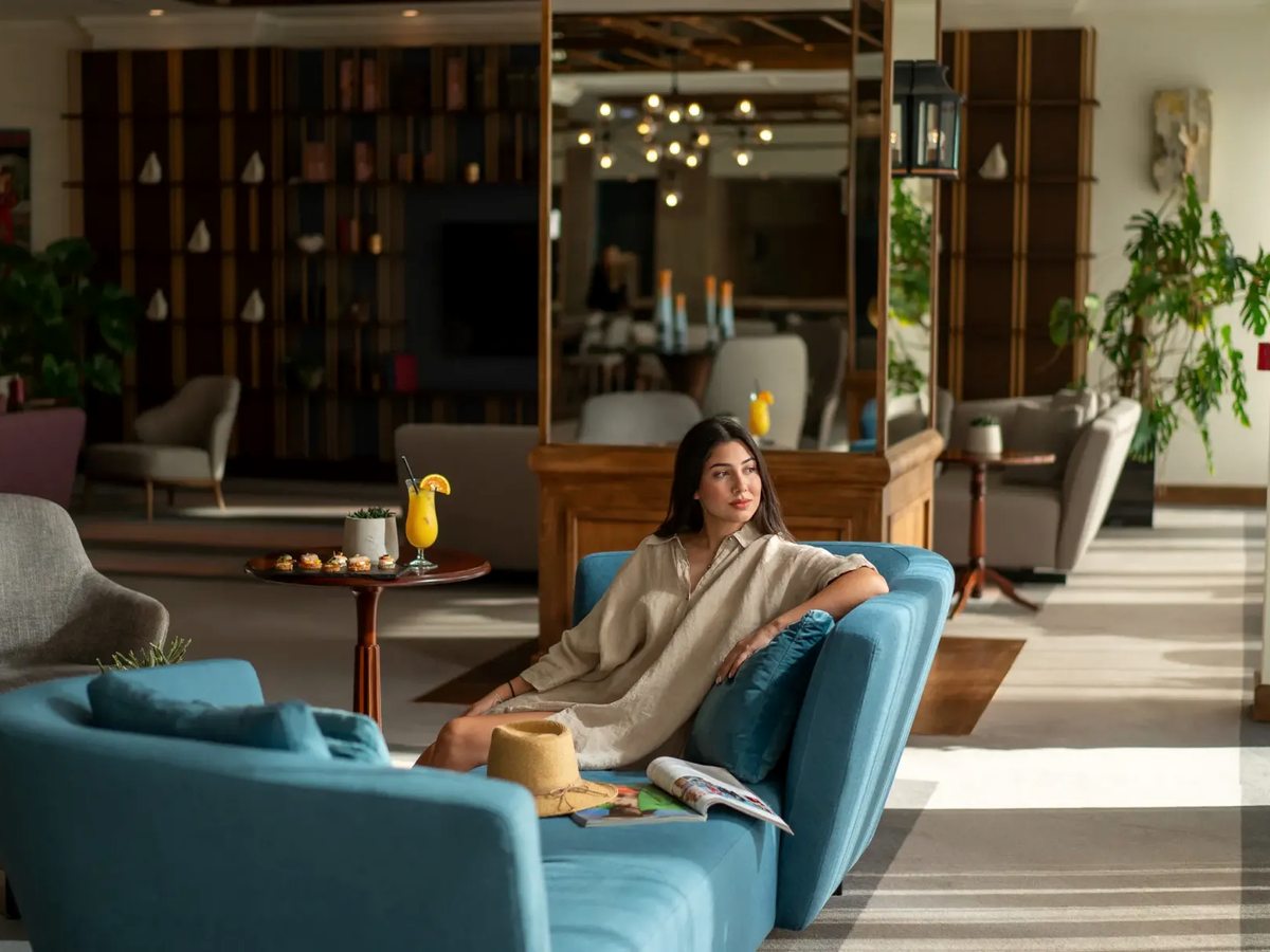 An open lobby lounge with turquoise sofas and grey armchairs around low tables, tall shelving and pendant lights, a woman relaxing on a sofa.