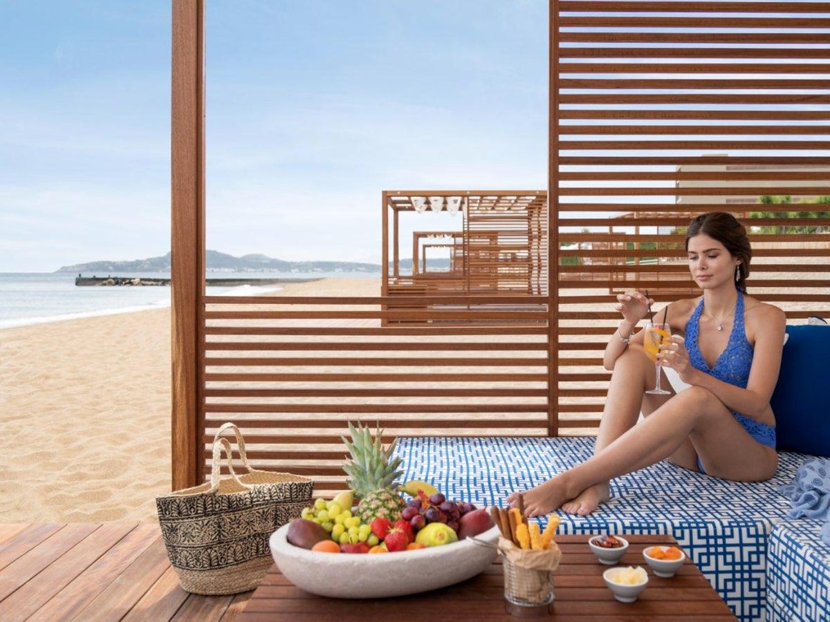 A slatted wooden beach cabana with a blue-patterned daybed, a bowl of fresh fruit on a low table and a woman with a drink, the beach and sea beyond.