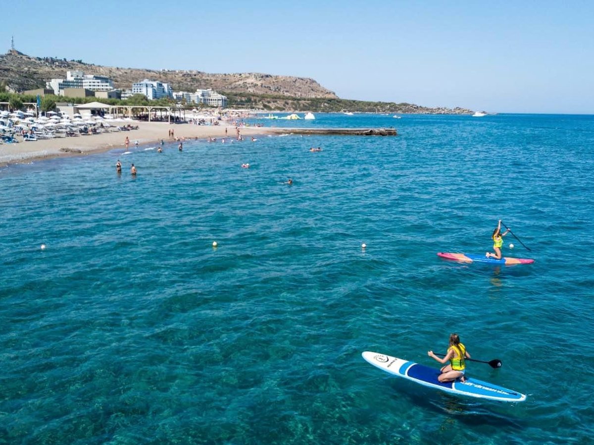 Two people paddleboard on clear turquoise water off the sandy resort beach, with swimmers near the shore and the coastline behind.
