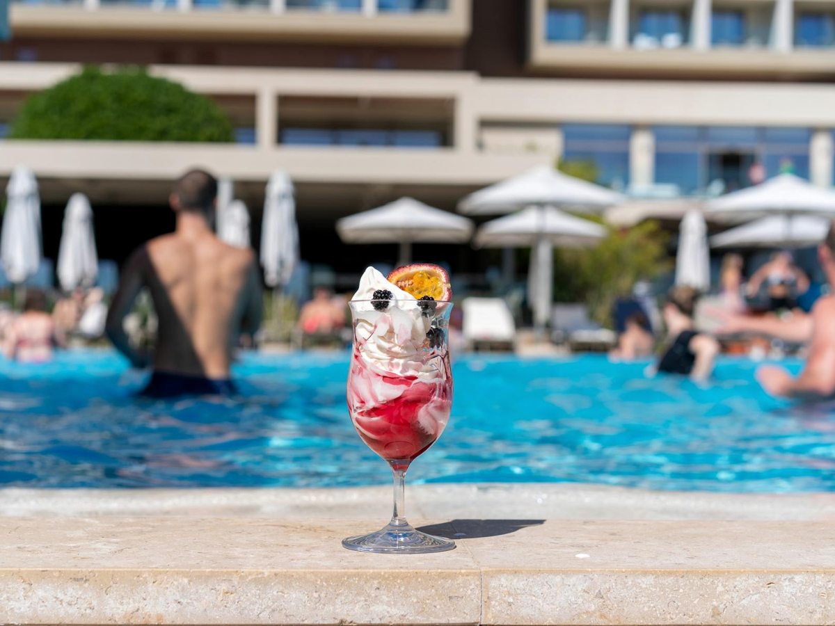 A tall fruit-topped dessert drink on the pool edge, with swimmers, umbrellas and the resort building blurred behind.