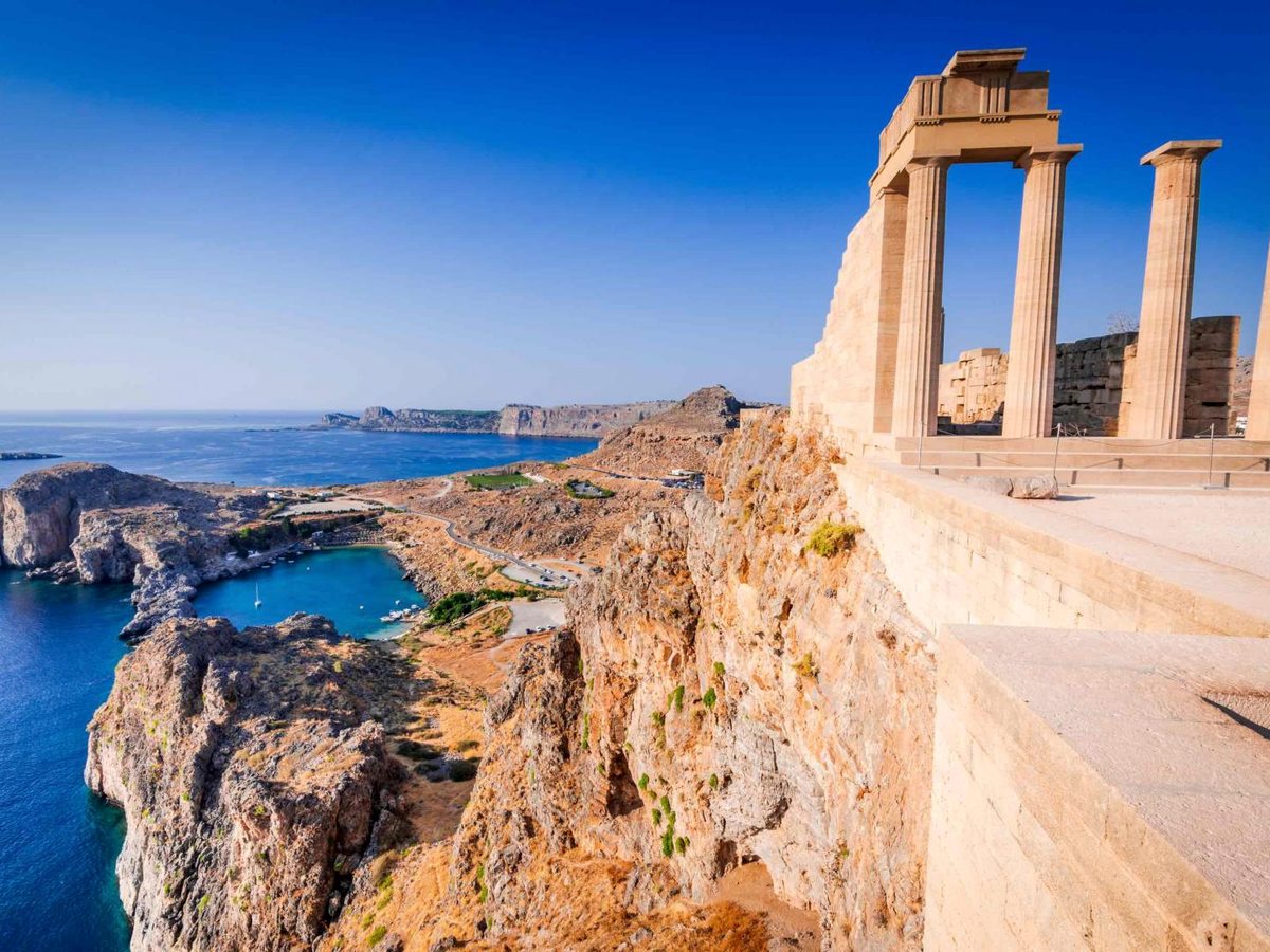 Ancient stone temple columns on a clifftop acropolis, with a sheltered blue bay and the coastline stretching away below.