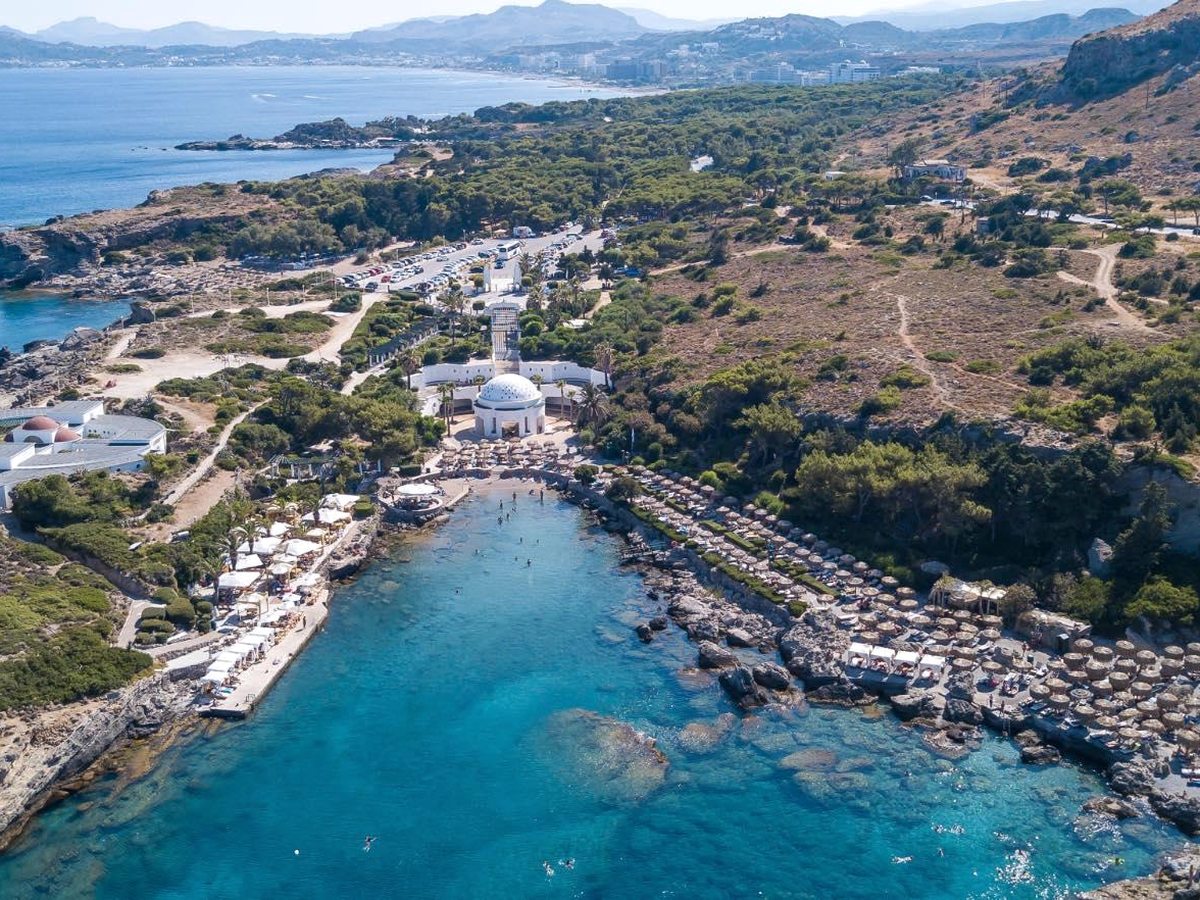 An aerial view of the domed white Kallithea Springs building above a small cove with rows of umbrellas and swimmers in clear turquoise water.