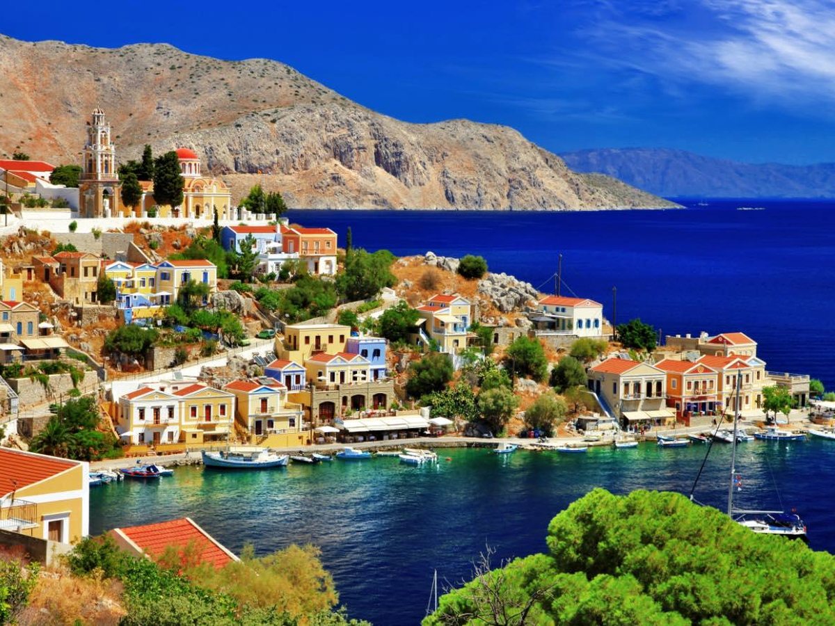 Pastel neoclassical houses with red-tiled roofs rising up a hillside around a small harbour full of boats, a bell tower on the ridge.