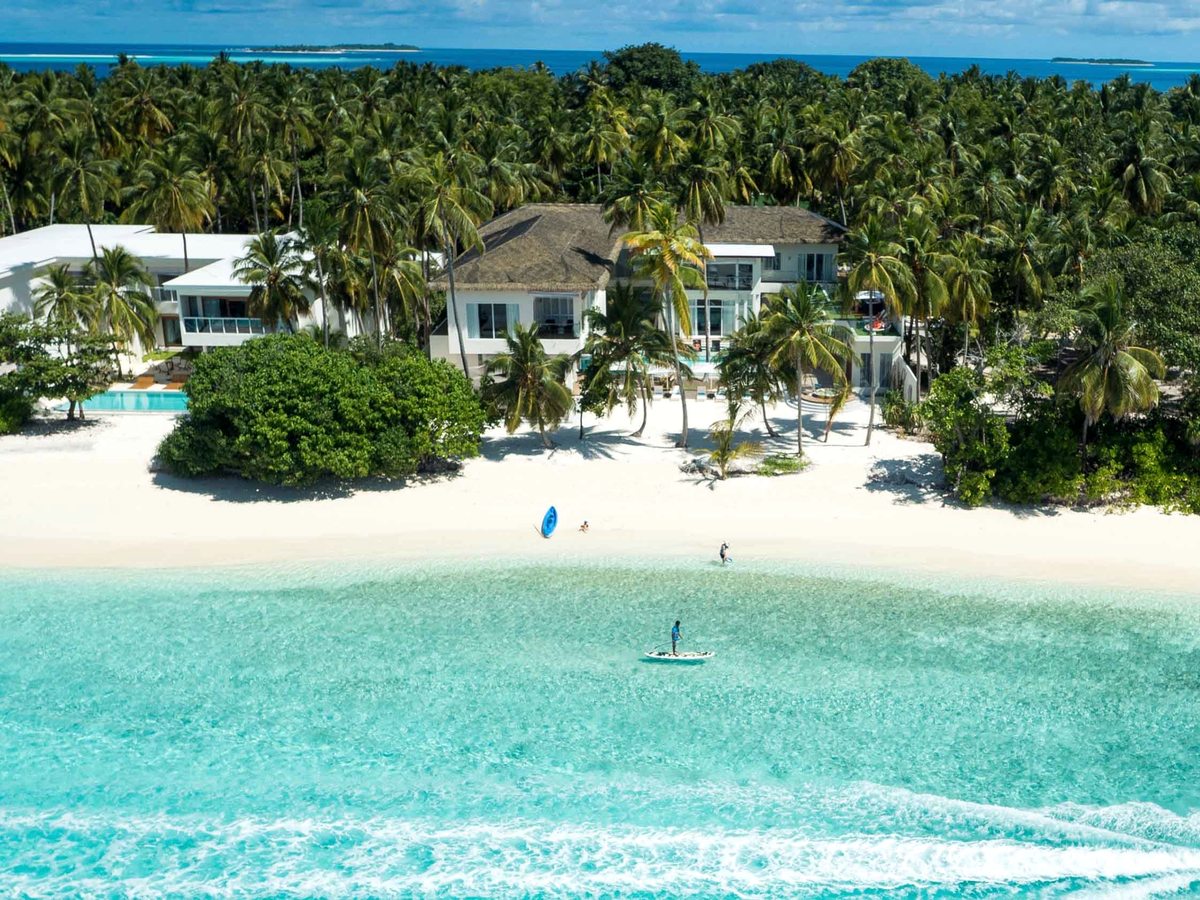 White sand beach with beachfront villas among palms, a calm turquoise lagoon and a stand-up paddleboarder in the water.