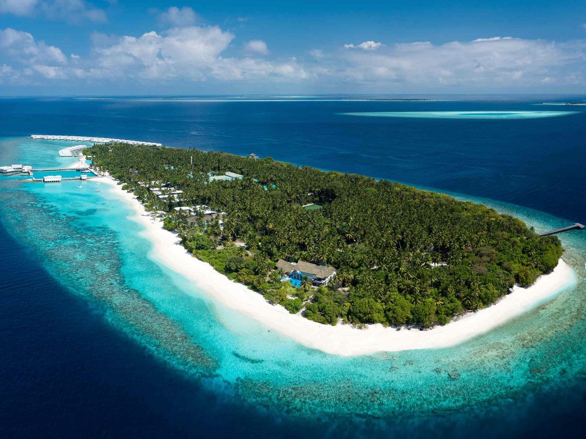 Aerial view of a long green jungle-covered island ringed by white sand and a turquoise lagoon, a line of overwater villas along one side.