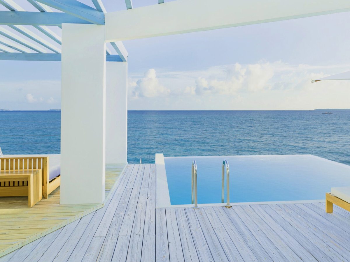 A timber villa deck over the sea with an infinity plunge pool, sun loungers and a shaded daybed looking out to open ocean.
